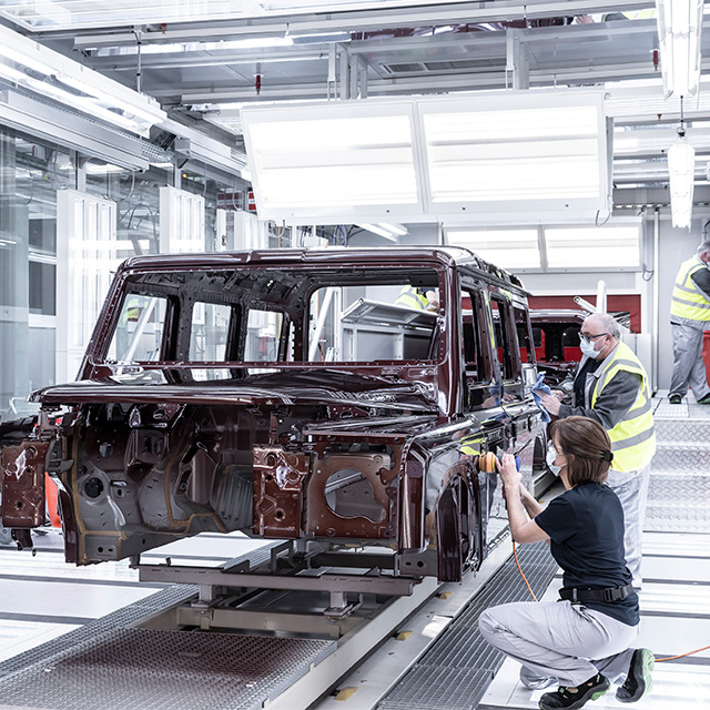 Photograph of a car manufacturing assembly line showing workers inspecting and assembling a dark red vehicle frame inside a brightly lit factory. The scene highlights detailed structural components of the vehicle body, with workers wearing safety vests and masks, emphasizing quality control and precision in automotive production.