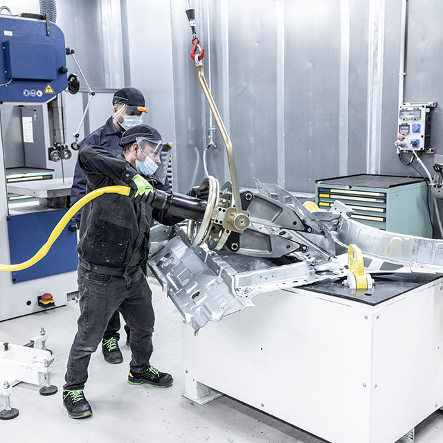 Photograph of two factory workers wearing masks and safety gear operating machinery to handle a large metallic automotive frame inside an industrial setting. The scene highlights use of a mechanical hoist with a yellow hose and a control panel in a clean, organized workspace focused on precision assembly or inspection.
