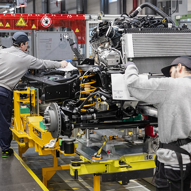 Photograph of two factory workers assembling a vehicle chassis and engine on a yellow production line platform inside an industrial facility. The scene highlights mechanical components like suspension springs, brake discs, and engine parts, with safety signs and equipment visible in the background.
