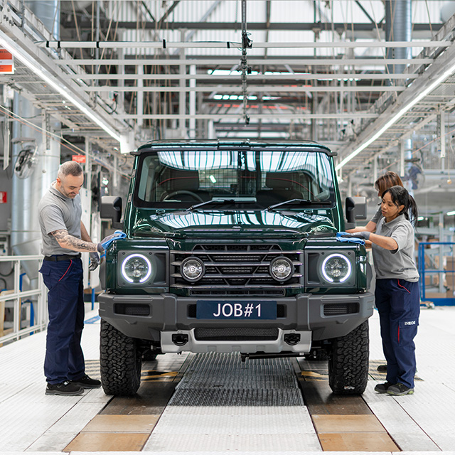 Photograph of a vehicle assembly line showing two workers inspecting a dark green off-road vehicle with illuminated round headlights and a license plate labeled "JOB#1." The factory setting features bright overhead lighting, metal framework, and a clean, organized workspace, highlighting a milestone in vehicle production or quality control.